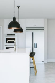 View of the kitchen island in a white kitchen. Dark lights over the island. Kitchen appliances in the background. Kitchen sink in island. 3D rendering.