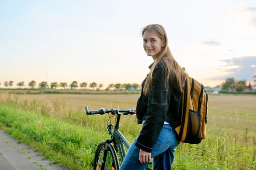 Teenage female with backpack on bike looking at camera, outdoor nature road sunset sky background, copyspace. Activity, lifestyle, youth concept