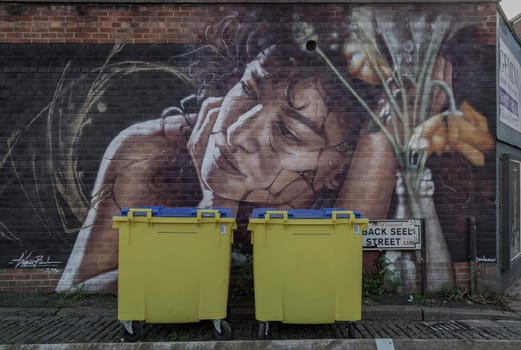 Liverpool, UK - Oct 09, 2023 - Two yellow Wheelie bin front of Portrait graffiti spray paint art pattern on the wall at Seel street, Space for text, Selective focus.