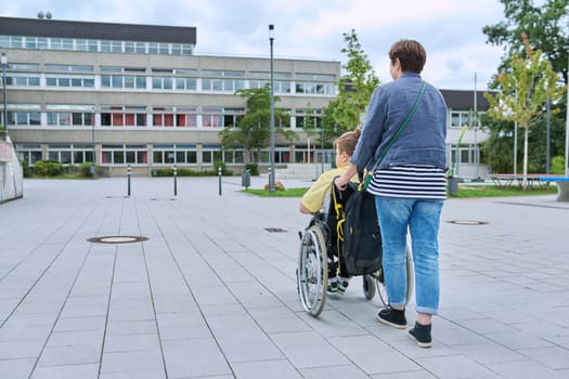 Back view of woman with boy in wheelchair near the school building, copy space. Children, disability, education, activity, health, lifestyle concept