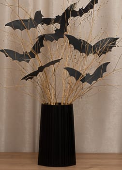 Halloween. Concept. Autumn holiday. Scary bats made of black paper, on dry branches in a black vase on the table. On a beige background. Close-up. Soft focus.