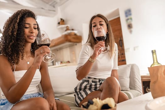 Two young women, different ethnicities, sit on the couch, sharing a blanket, laughing, enjoying winter at home with tea and pastries.