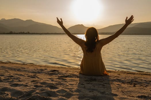 Evocative shot: woman in contemplative pose sitting on the sand with arms wide, facing the sea at sunrise or sunset on a beach. Connect with nature.