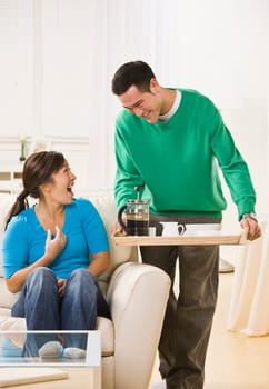 A man standing with coffee service and a woman sitting down with a surprised look on her face. Vertically framed photo.