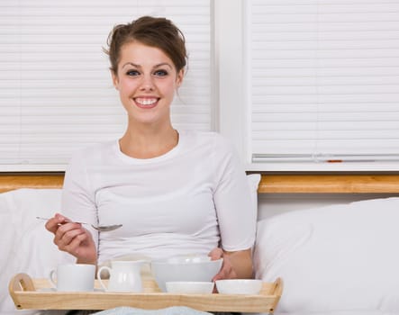 An attractive young woman eating breakfast in bed. She is smiling at the camera. Horizontally framed shot.