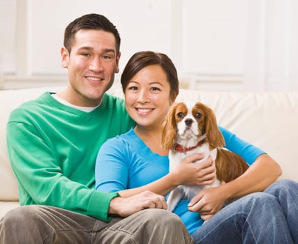 An attractive young asian couple sitting on a couch together and holding a dog. They are smiling at the camera. Horizontally framed shot.