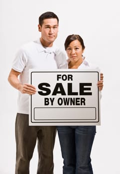 A worried looking asian couple holding a 'For Sale' sign. They are looking directly at the camera. Vertically framed shot.