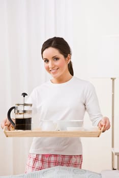 A young, attractive woman is standing in her pajamas and is holding a breakfast tray. She is smiling at the camera. Vertically framed shot.