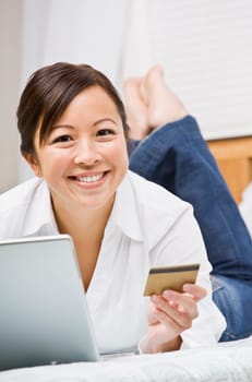 Beautiful asian woman holding a credit card and using a laptop. She is smiling at the camera. Vertically framed shot.