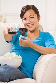 A beautiful young asian woman sitting on a couch. She is holding a bowl of popcorn and is using a remote. She is smiling directly at the camera. Vertically framed shot.