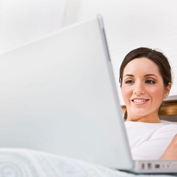 A beautiful young woman is sitting in a room and working on a laptop. She is smiling and is looking at the screen. Square framed shot.