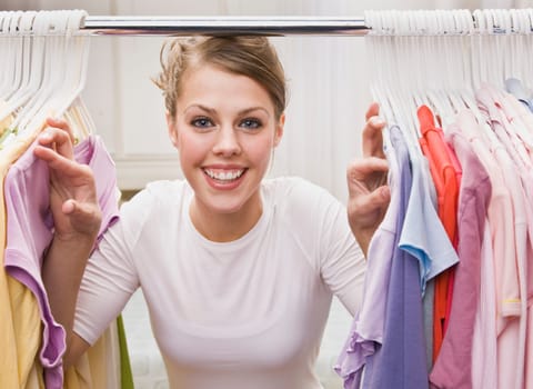 A young, attractive woman is standing in a closet and looking through clothes. She is smiling at the camera. Horizontally framed shot.