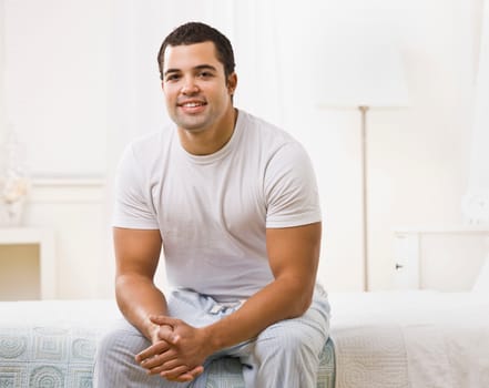 A happy looking man seated on a bed in a white room. He is smiling and looking at the camera. Horizontally framed shot.