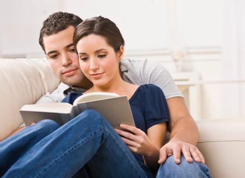 A young, attractive couple is seated together on a couch and are reading a book together. They are looking away from the camera. Horizontally framed shot.