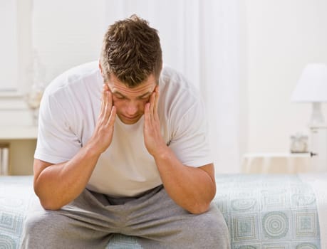 A man is seated on the bed in a bedroom. He has his head in his hands and is looking away from the camera. Horizontally framed shot.