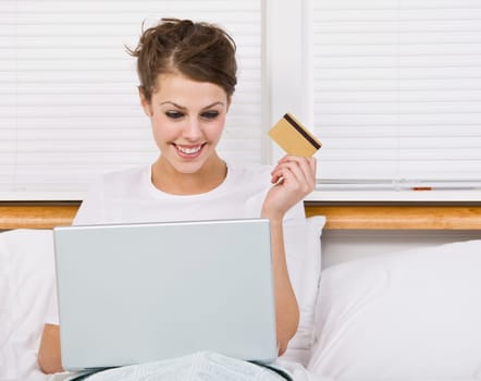 An attractive young woman holding a credit card while using a laptop. She is smiling at the laptop screen. Horizontally framed shot.