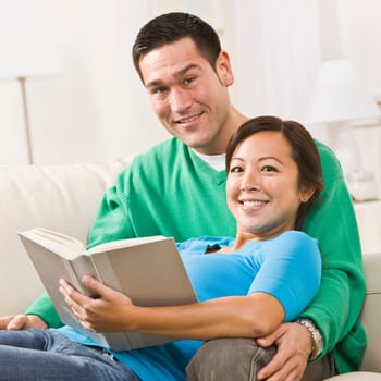An attractive young couple sitting on a couch together with a book. The female is sitting in the male's lap. They are smiling directly at the camera. Square framed shot.