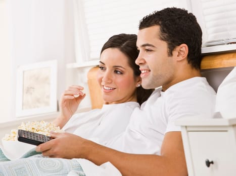 An attractive young couple lying in bed and watching television. They have a bowl of popcorn and a remote. They are smiling. Horizontally framed photo.