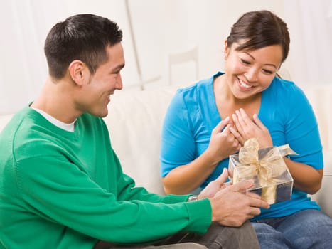 An attractive young couple seated on a couch. The male is presenting a gift to the female. They are both smiling. Horizontally framed shot.