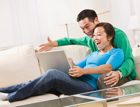 An attractive young couple sitting on a couch together and viewing laptop screen. They are laughing. Horizontally framed shot.