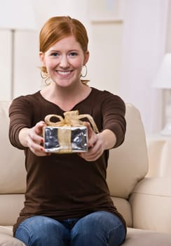 An attractive young female sitting on a couch and presenting a gift with outstretched arms. She is looking directly at the camera and is smiling. Vertically framed photo.