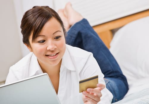 An attractive young woman lying on a bed and gazing at a credit card. She is holding a laptop and is smiling. Horizontally framed photo.