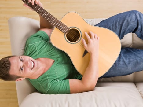 An above view of an attractive young man lying on the couch and playing a guitar. He is smiling directly at the camera. Horizontally framed shot.