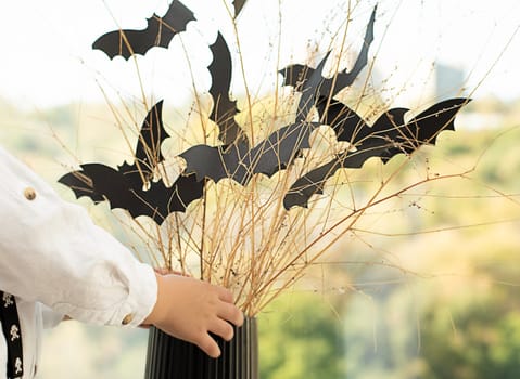 Halloween concept. No face. The baby's hands near the window, against the background of a vase with dry branches and black paper bats. Close-up. Soft focus. Background.