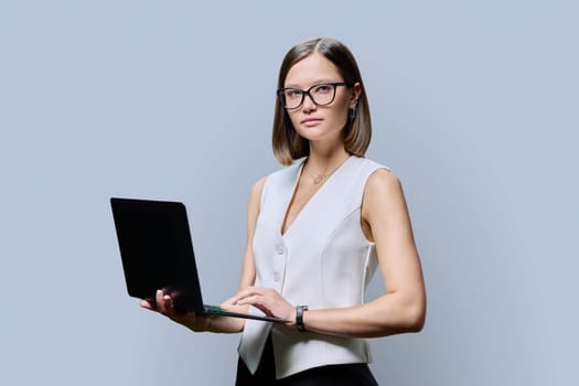 Serious young business woman with laptop on grey studio background. Successful confident caucasian female looking at camera. Business, work, job, study, education, e-learning, technology concept