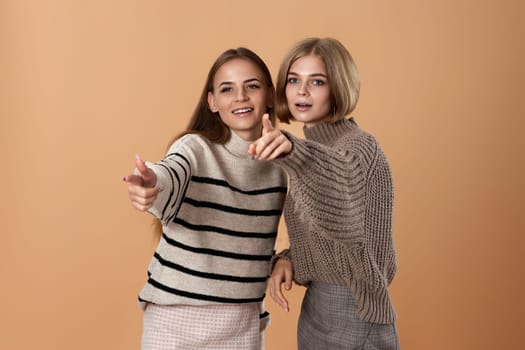 Two women friends together in casual clothes point index finger aside on beige background. Surprised blonde woman and her friend