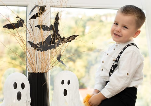 Halloween. Concept. A cute and cheerful boy sits near the window against the background of white ghosts and a vase with dry branches and black paper bats. Close-up.