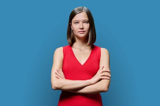 Portrait of young confident woman in red on blue studio background. Successful fashionable female with crossed arms looking at camera. Business, work, services, education, fashion beauty professions