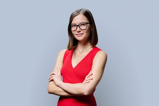 Young confident woman in glasses in red with crossed arms looking at camera on grey studio background. Business, work, services, education, fashion, beauty, professions, people