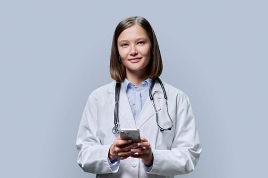 Young woman doctor, medical student intern with smartphone in hands on grey studio background. Technology, medicine, online Internet communications, medical app services, telemedicine