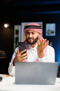 Arab guy engrossed in a video conference call with a coworker on his smartphone. He expertly uses wireless technology for work and research, showcasing his digital communication skills.