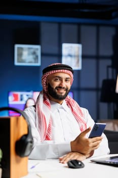 With a laptop nearby, a Muslim man holds a mobile device and confidently looks at the camera. The Arab tech-savvy entrepreneur combines digital and traditional aspects of his career.