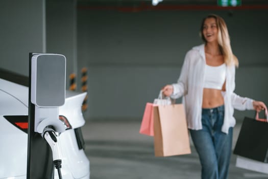 Young woman travel with EV electric car to shopping center parking lot charging in downtown city showing urban sustainability lifestyle by green clean rechargeable energy of electric vehicle innards