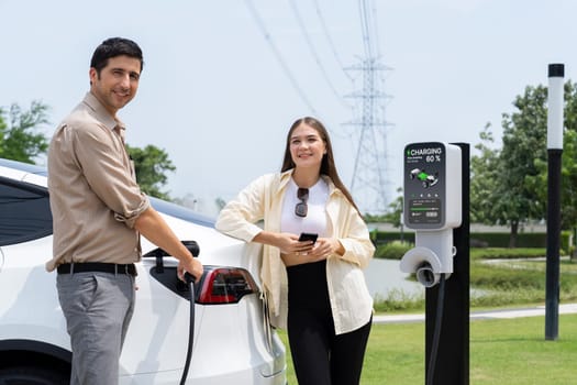 Young couple use smartphone to pay for electricity at public EV car charging station green city park. Modern environmental and sustainable urban lifestyle with EV vehicle. Expedient