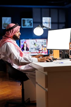 Image shows an Islamic man sitting at his home office desk with the desktop pc monitor displaying a blank copyspace template. Dedicated Arab guy using the computer having an isolated white screen.