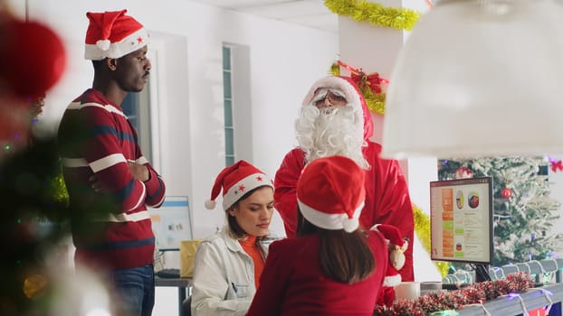 Revealing shot of manager dressed as Santa during Christmas season delegates project tasks to workers. Management executive in xmas decorated workspace discussing with employees