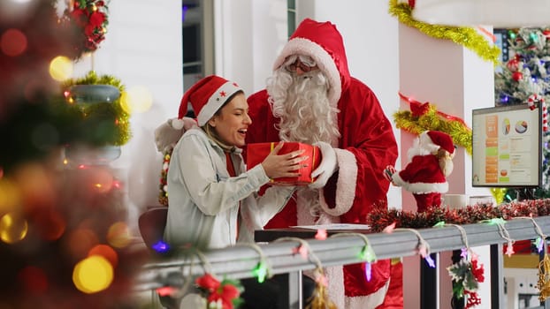 Revealing shot of worker receiving gifts from colleague dressed as Santa Claus in xmas ornate workspace. Disguised coworker surprising woman with presents before Christmas office celebrations