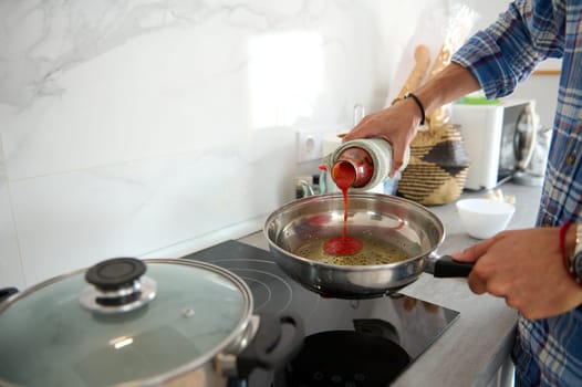 Details on male chef hand holding a bottle with passata and adding tomato sauce into a frying pan with olive oil and bay leaf, cooking pasta for dinner, according Italian traditional family recipe