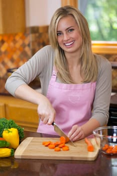 A young woman is slicing carrots in her kitchen and smiling at the camera.  Vertically framed shot.
