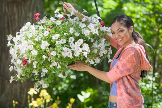 A young woman smiles while posing next to a hanging basket of flowers. Horizontal format.