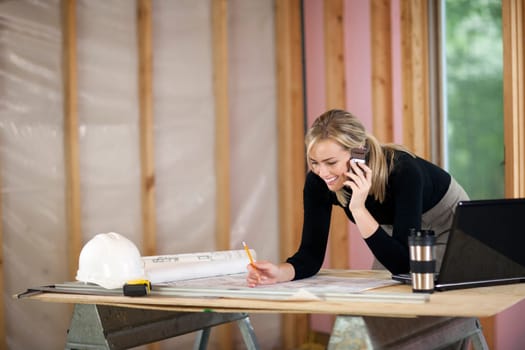 A young woman is looking at blueprints and talking on the phone.  She is working at a construction site.  Horizontally framed shot.