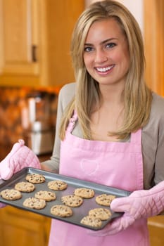 A young woman is holding a baking sheet of cookies and smiling at the camera.  Vertically framed shot.