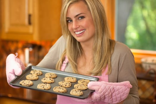 A young woman is holding a baking sheet of cookies and smiling at the camera.  Horizontally framed shot.