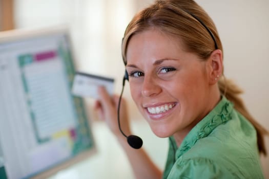Close-up of a smiling young woman in front of a computer, wearing a headset and holding a credit card. Horizontal format.