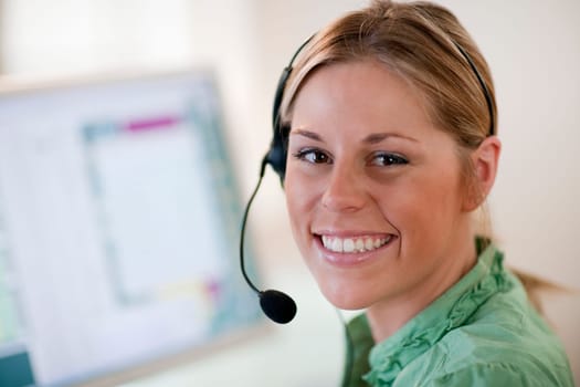 Close-up of a young woman in front of a computer, wearing a headset and smiling at the camera. Horizontal format.