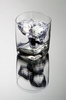 Empty glass tumbler with ice cubes on grey background with reflection